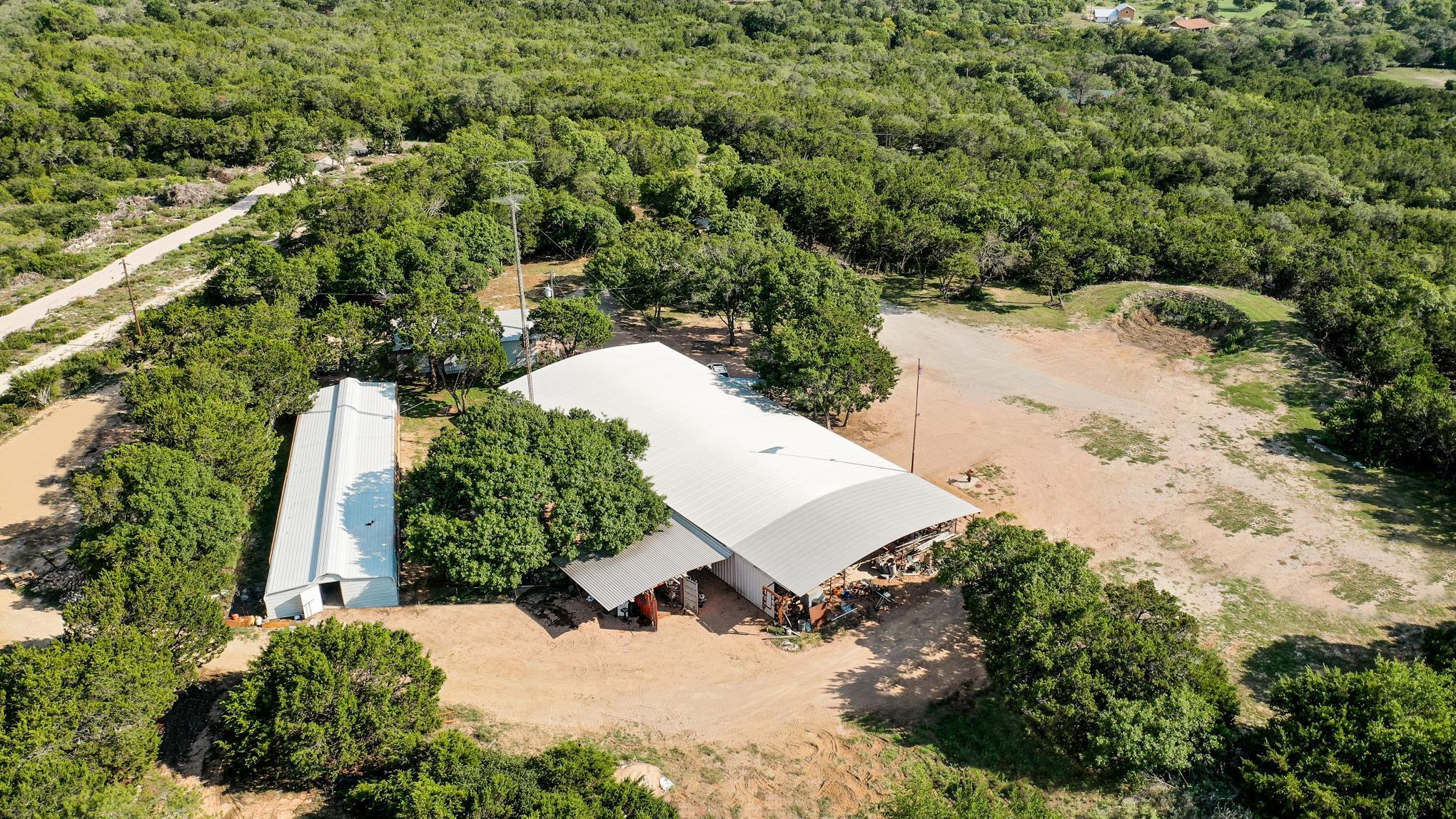 REDUCED $50K! HECK OF A DEAL for a business owner or family compound. Resting on just over 9 private, wooded acres, 2 miles from Burnet, and conveniently located off Hwy 29, this 6,000 square foot bardominium “WORKS.” Combining home and business effortlessly in one convenient building, each run separate meters, 200-amp service, and both are 3,000 square feet. From the entry, step into the large living and dining rooms with a built-in entertainment center and two ceiling fans. The convenient galley kitchen is efficient with granite counters and built-in appliances while the owner suite provides a built-in entertainment center, an on-suite bath, walk-in closet, and convenient access to the laundry closet. A large second bedroom shares the laundry closet, and toward the front of the house, a flex room could serve as an office, craft room, bunk room, or extra storage space. Across from the living room, an oversized third bedroom furnishes additional cabinetry and a convenient half bath. Outdoors, live oaks shade the backyard and the barn adds convenient storage. A FEMA-style trailer with 50-amp service, grinder pumps, and dedicated washer/dryer hookups provide further accommodation. Shop has two double sliding garage doors and 3 roll up doors. Septic with 1500 gallon holding tank & pump south of the shop (new pump installed 3 yrs. ago); drain field west of the house: 40’x48’. RV hookup next to the well house (well 280’ deep) offers 50-amp hookup. House has two 50-gal hot water heaters, installed in-line & 4-z